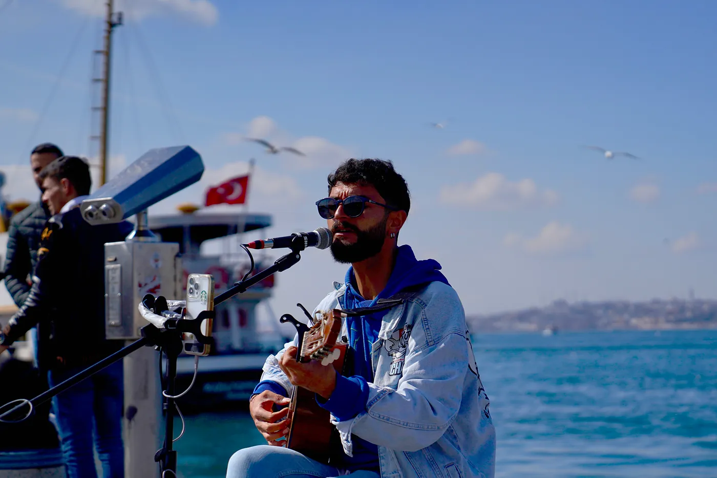A street musician playing his guitar at Kadıköy's waterfront with the district of Fatih across the Bosphorus.