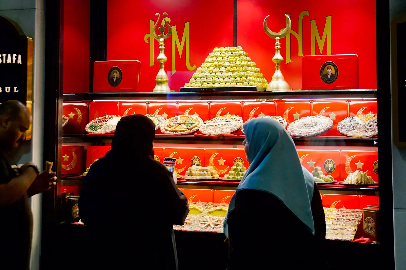 Onlookers inspecting the desserts of a Hazır Mustafa shop at sundown.