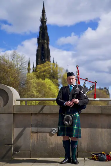A bagpiper in full Highland dress playing in Princes Street Gardens, the Scott Monument behind him